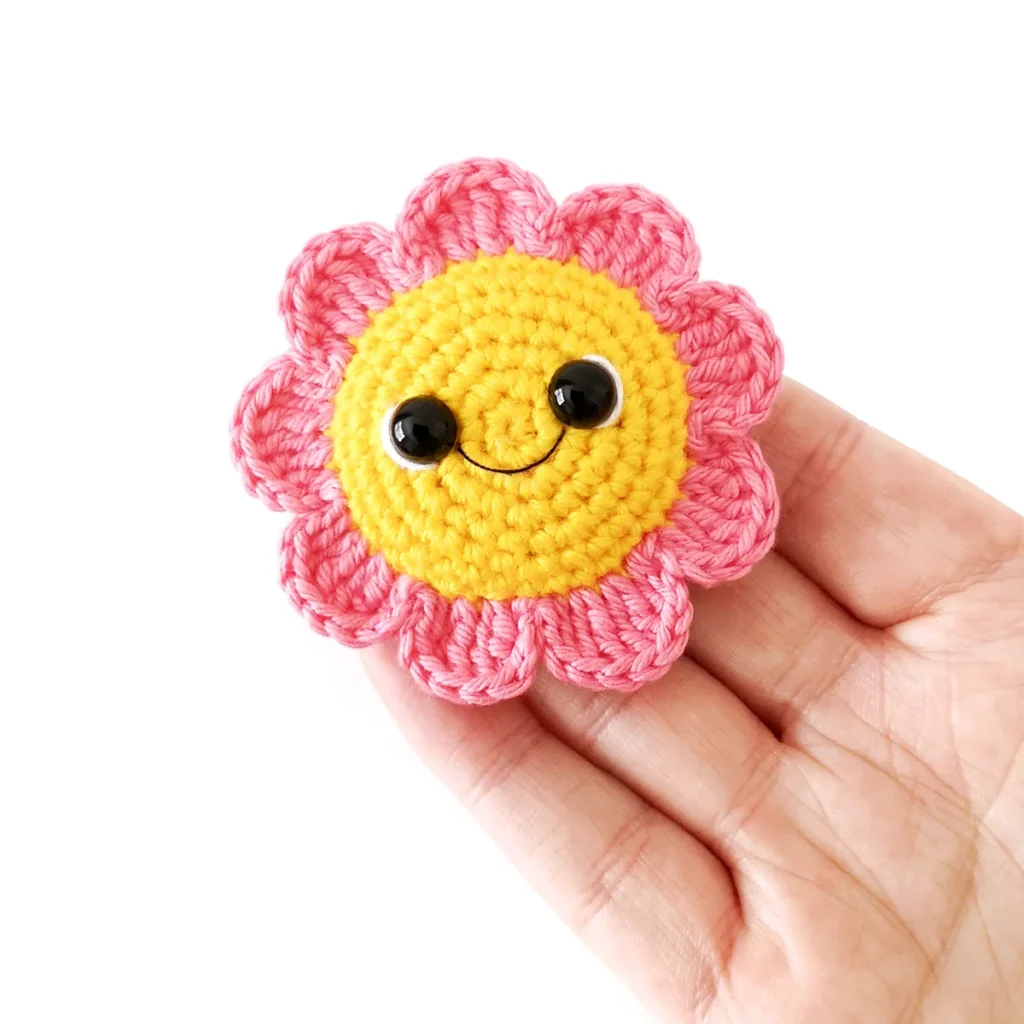 A pink crochet amigurumi flower with yellow centre and smiling face, lying flat on the end of a hand against a white background.