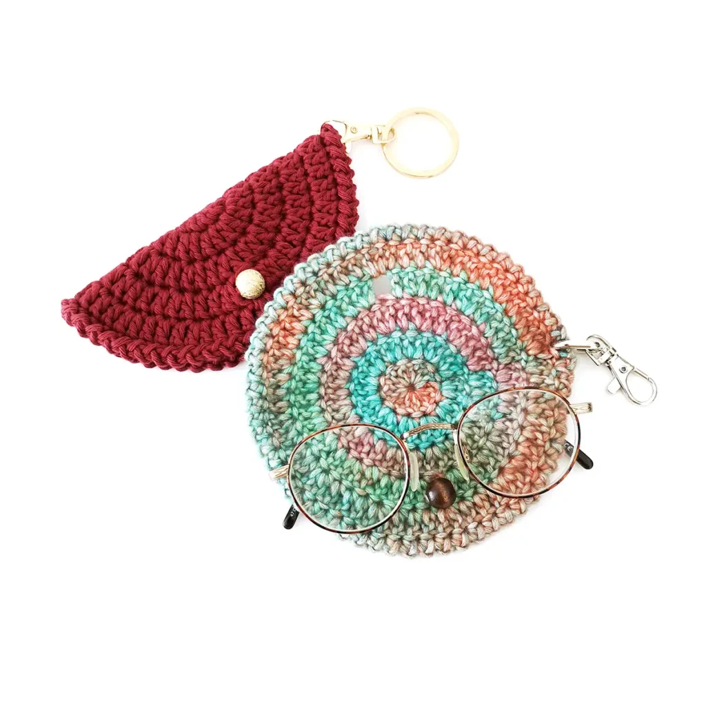 Two crochet glasses cases, one open and slightly overlapping, with gold keychain clasps and round bead closures, and a pair of glasses lying on top of the front case.