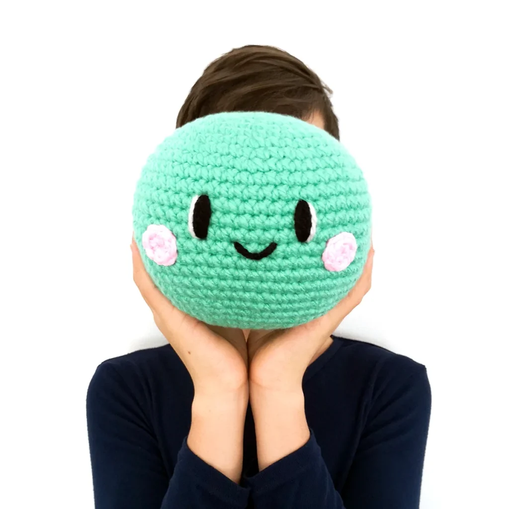 A single crochet pea with a smiling face and pink crocheted cheeks, being held up in front of a persons face.
