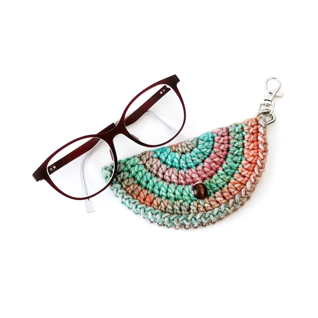 A multicoloured crochet glasses case with silver lobster clasp and round brown bead closure, and a pair of glasses against a white background.