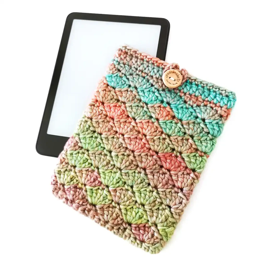A crochet kindle cover lying overlapping a kindle slightly, featuring the shell stitch and made with soft gradient yarn in shades of green, blue, orange and purple, with a round wooden button and loop closure.