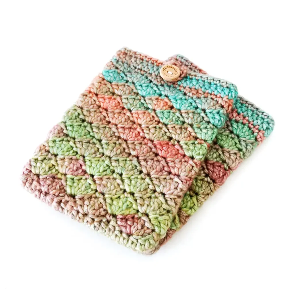 Two crochet kindle covers lying stacked on top of each other, featuring the shell stitch and made with soft gradient yarn in shades of green, blue, orange and purple, with a round wooden button and loop closure.