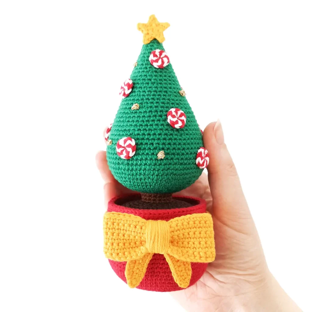 Crochet Christmas tree featuring a red pot, large yellow crochet bow and star, decorated with red and white crocheted peppermints and gold French knots being held up against a white background.