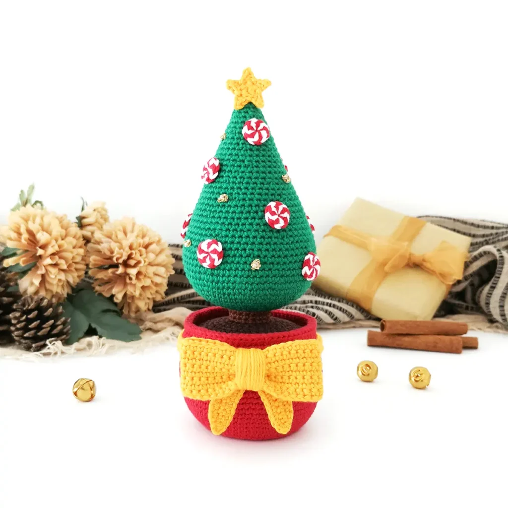 Crochet Christmas tree featuring a red pot, large yellow crochet bow and star, decorated with red and white crocheted peppermints and gold French knots with gold bells, cinnamon sticks, pinecones, fake flowers and a black and beige striped scarf in the background.