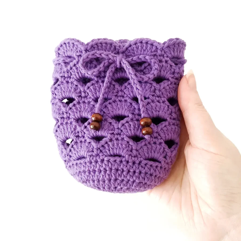 A purple iced coffee cozy crochet pattern with beaded tie tied in a bow, being held up by a hand against a white background.