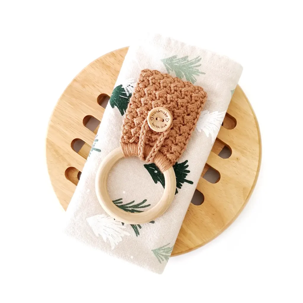 A brown crochet towel holder with button loop closure and wooden ring, lying on a folded kitchen towel and round wooden pot stand.