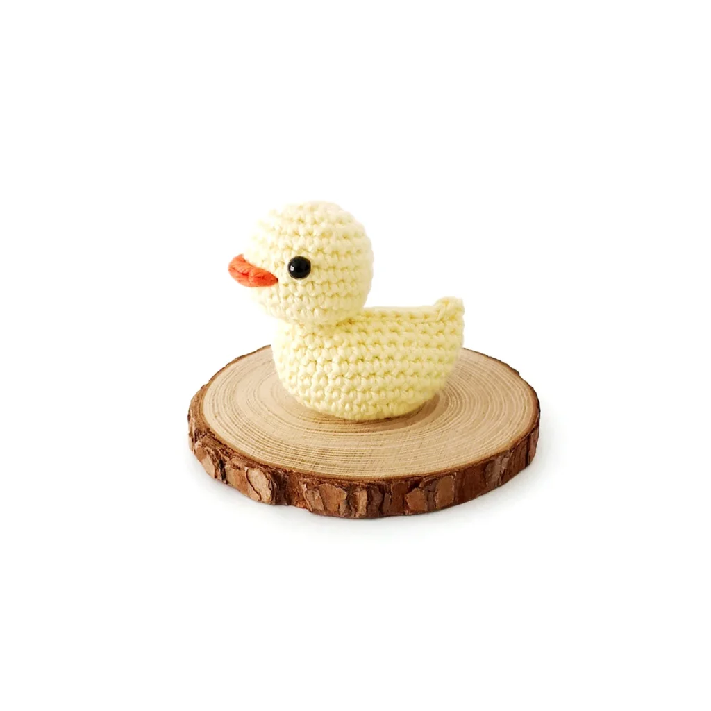 A little yellow crochet duck with small black eyes and an orange beak, sitting on a wood round.