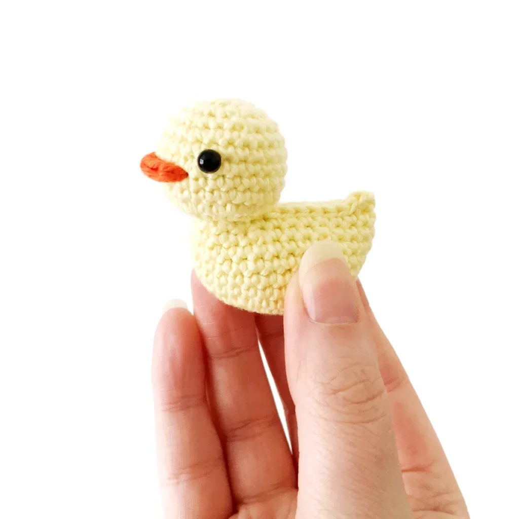 A little yellow crochet duck with small black eyes and an orange beak, being held up by a hand, against a white background.