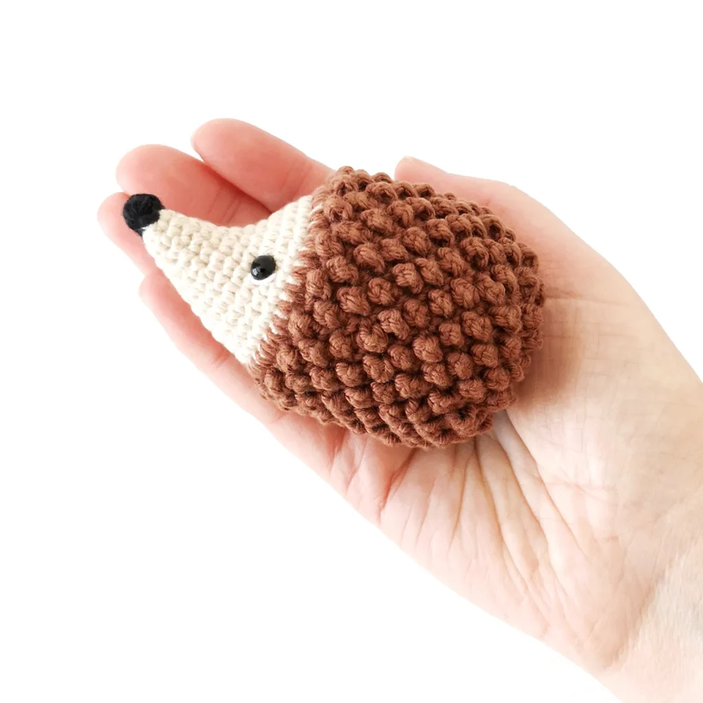 A crochet hedgehog with small black nose and eyes, held in the palm of a hand.