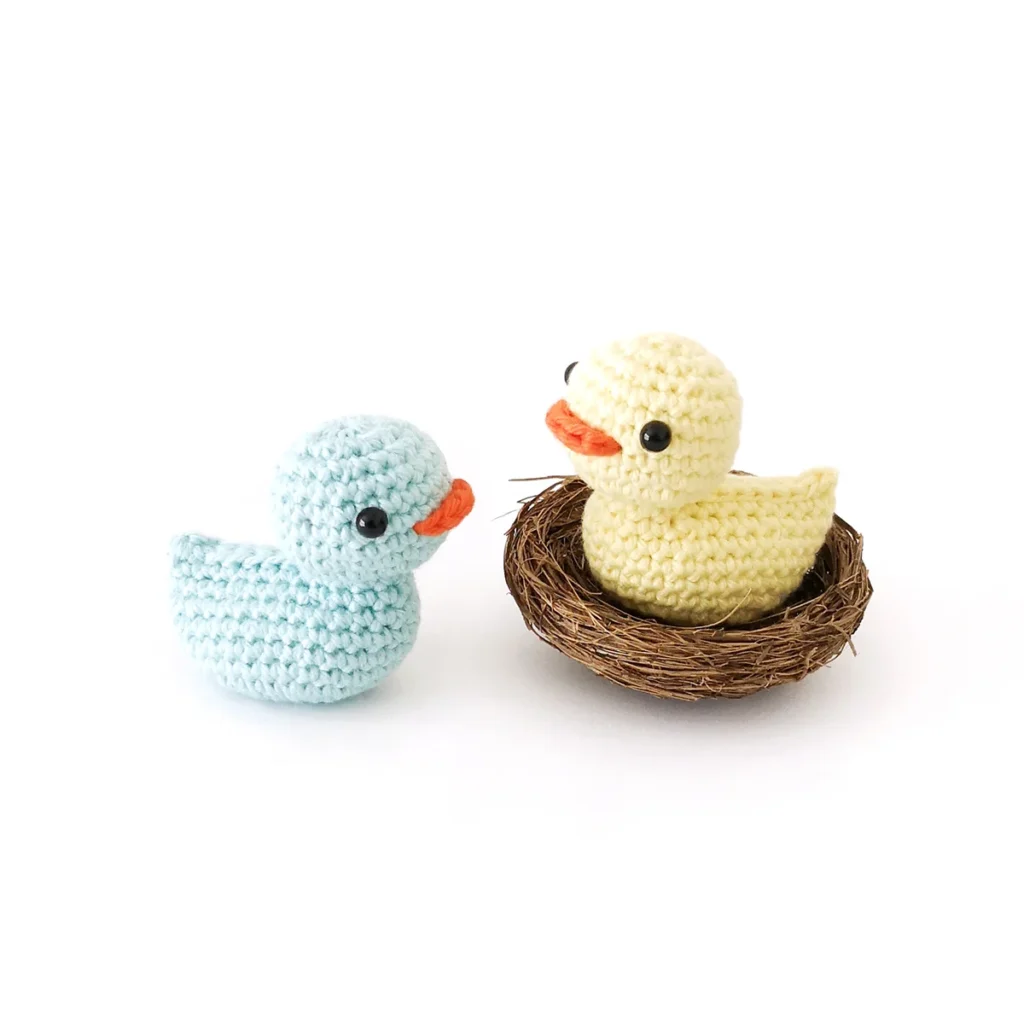 A little yellow crochet duck with small black eyes and an orange beak sitting in a brown twig nest with a light blue crocheted duck next to it.