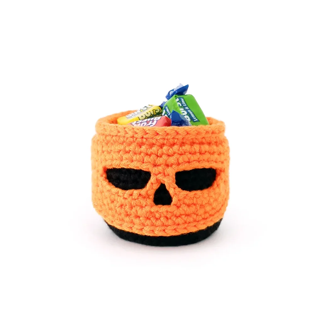 A small orange and black pumpkin skull basket filled with wrapped candy.