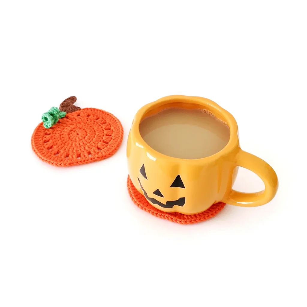 An orange crochet pumpkin coaster with a brown stem and small green vine, lying next to an orange jack-o'-lantern mug.