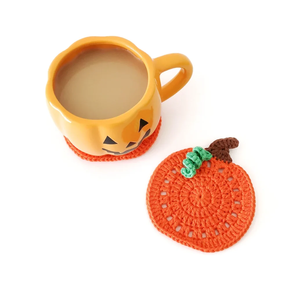 An orange crochet pumpkin coaster with a brown stem and small green vine, lying next to an orange jack-o'-lantern mug.