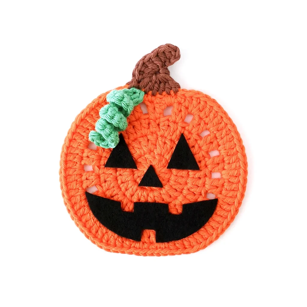 An orange crochet pumpkin coaster with brown stem and small green vine, with a jack-o'-lantern face.