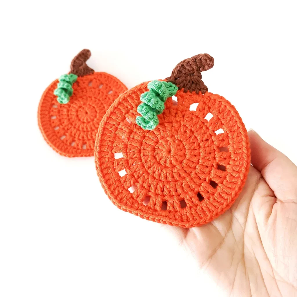 Two crochet pumpkin coasters with brown stems and small green vines, with one being held up to the camera.